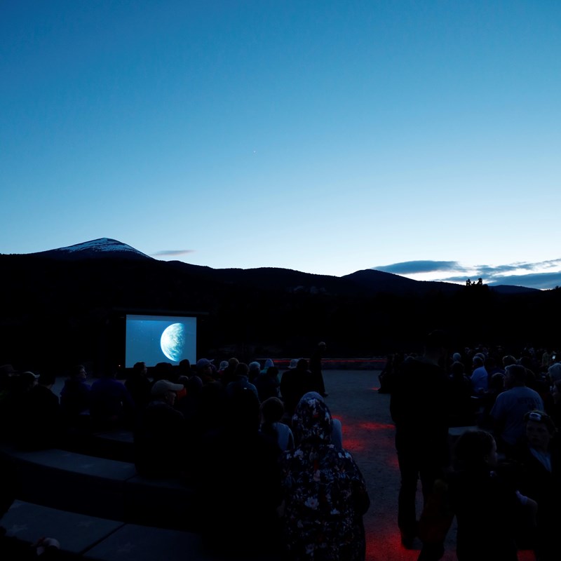 A crowd sits in a darkened amphitheater before a lit up screen and mountains in the distance. Dusk.