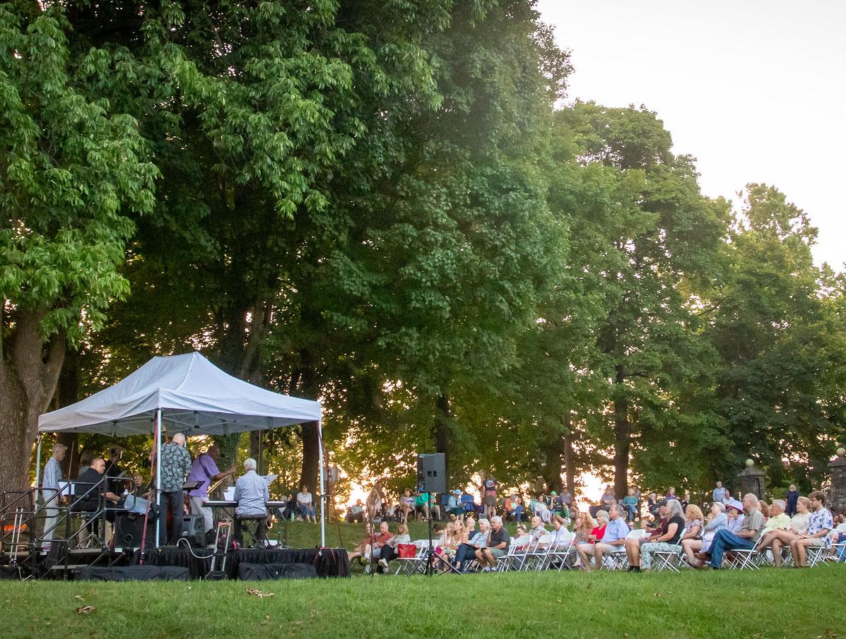 Musicians play under a tent in front of a crowd on a lawn, surrounded by large trees.