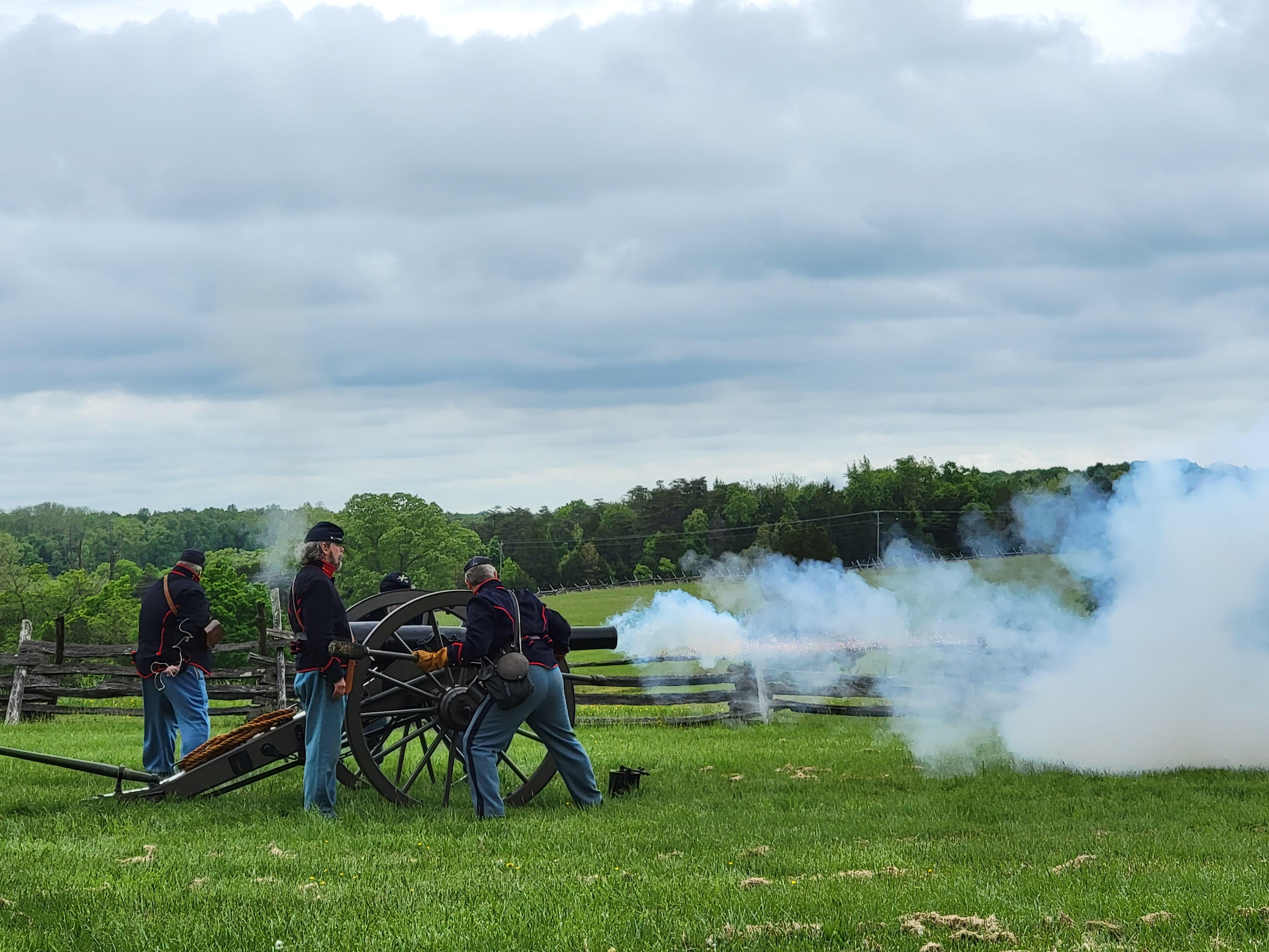Men in living history attire fire a cannon in a green field. A stream of smoke surrounds them.
