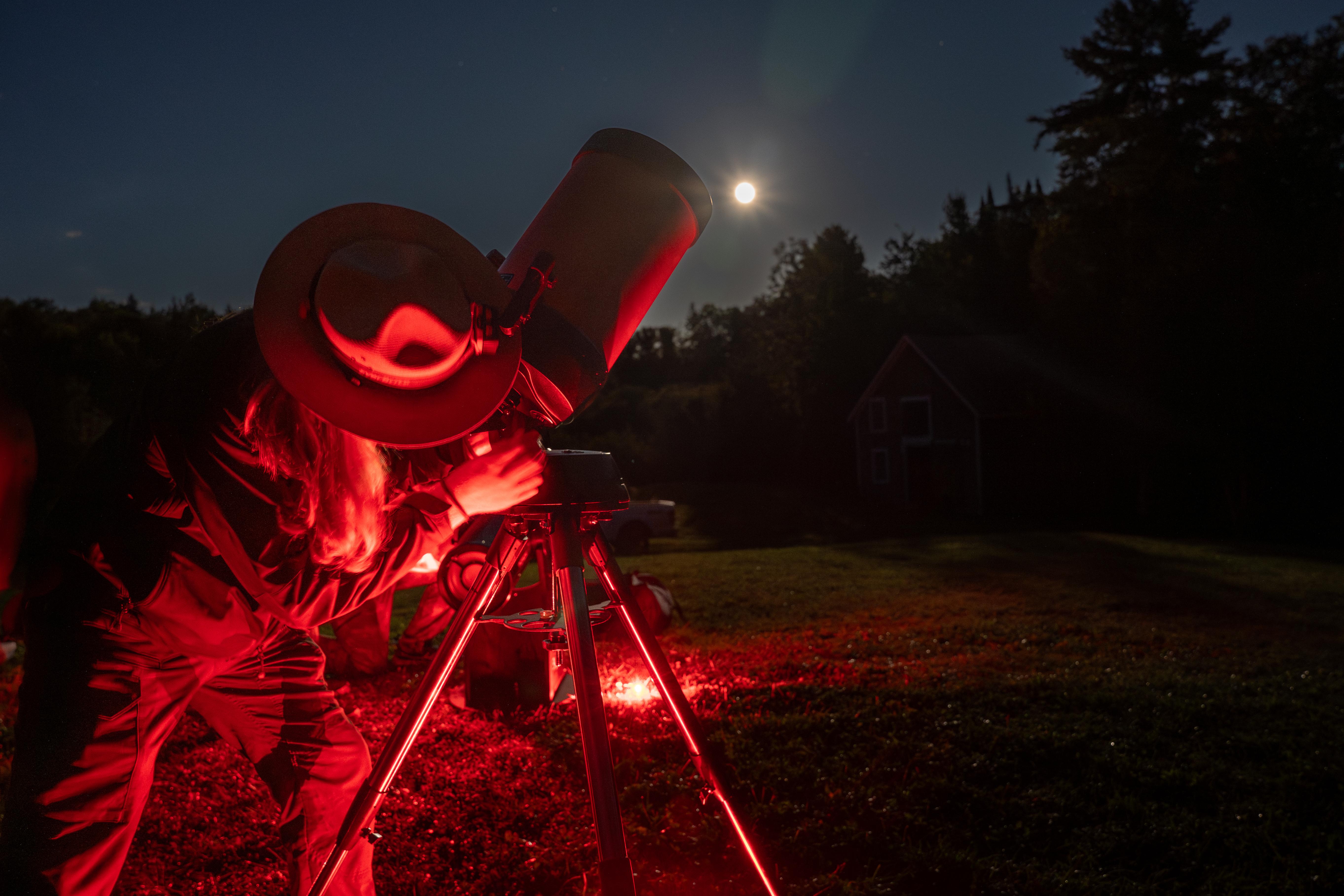 A ranger peers through a telescope with the moon overhead in the background.