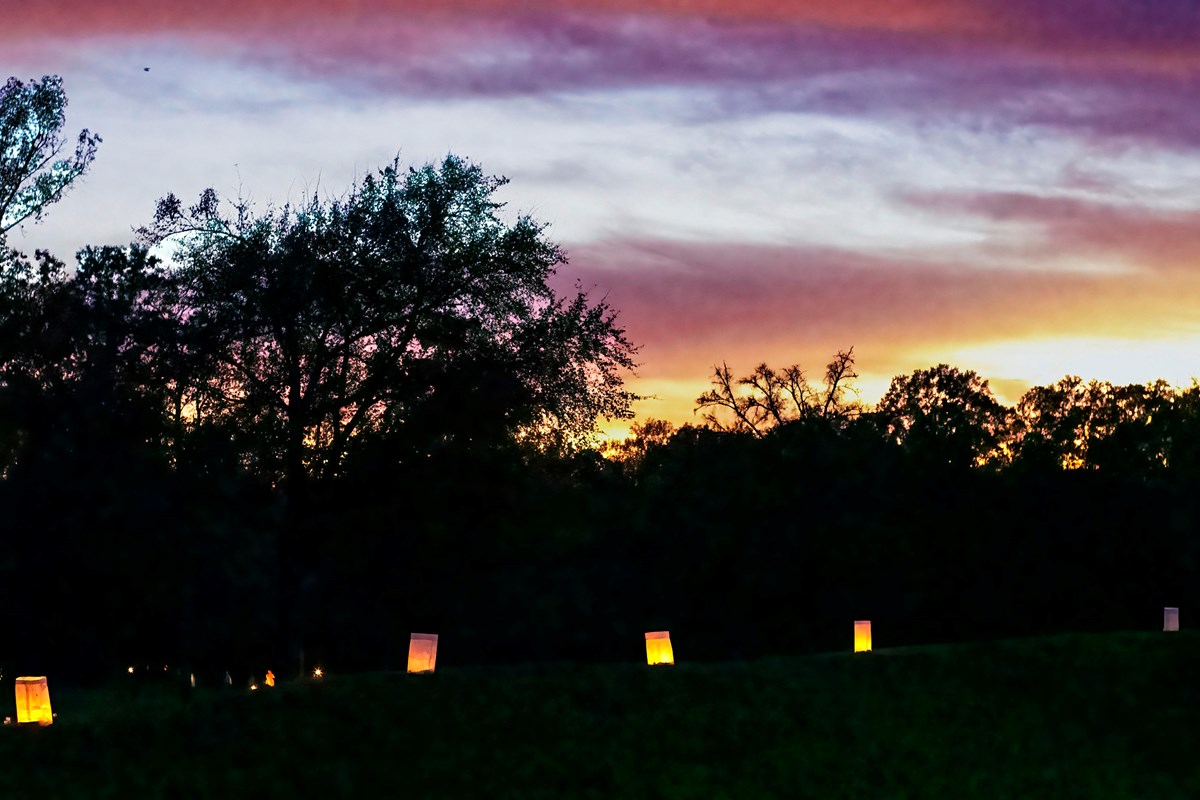 A purple and yellow sunset behind trees with luminaries litup in the forground.