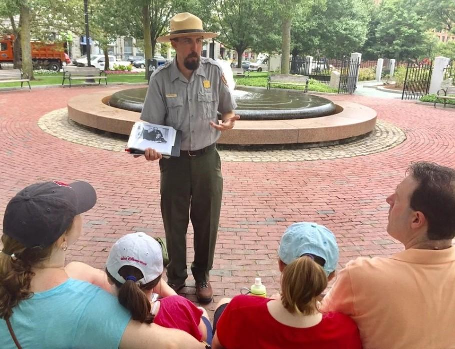 A uniformed park ranger holds a photo and speaks to a family gathered in front.