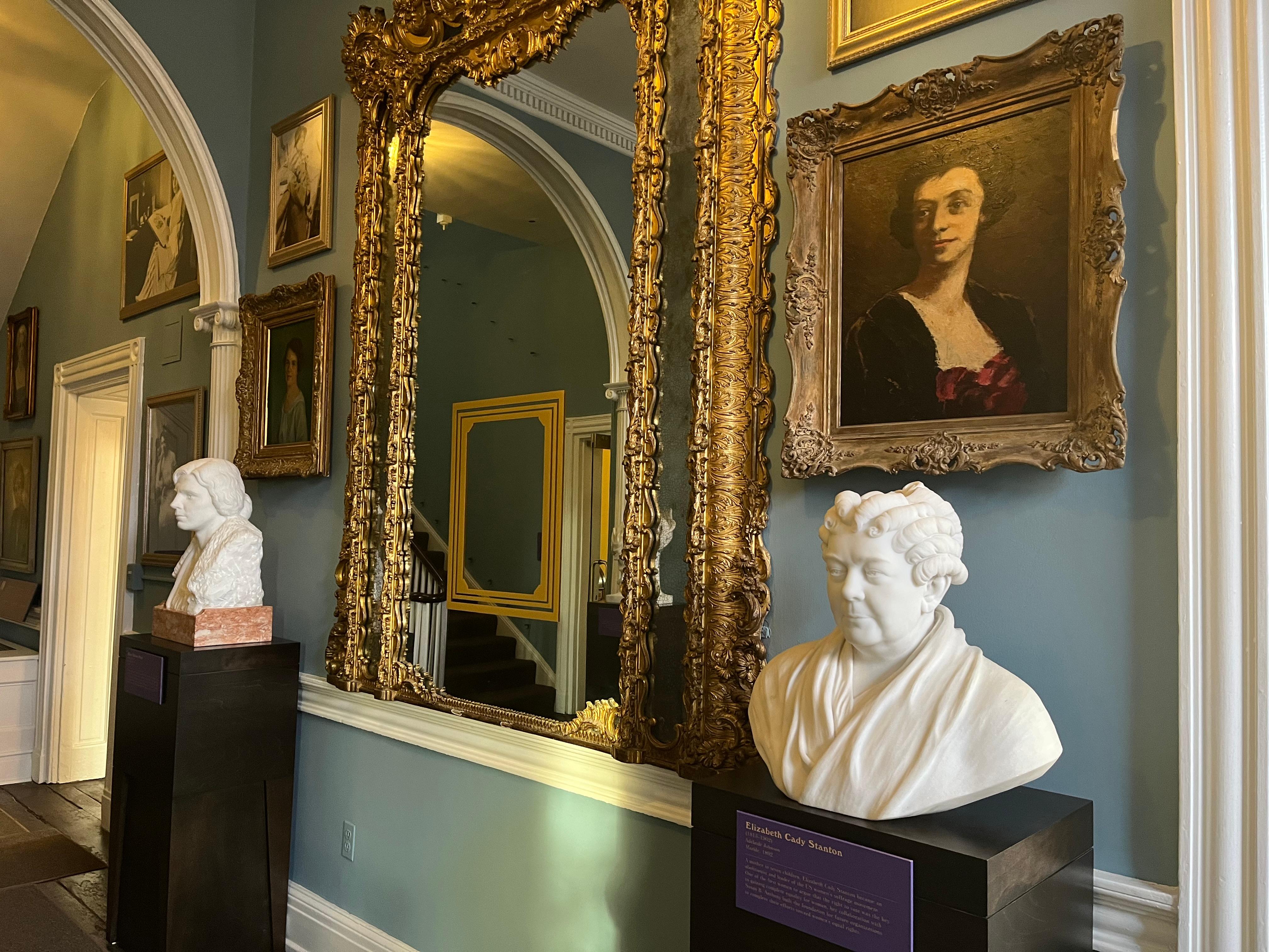 Hallway with large mirror in center surrounded by portraits on walls and busts on pedestals