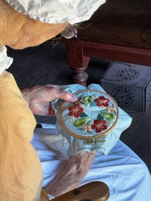 A volunteer in colonial dress practices cross stich. She is stitching a flower design.