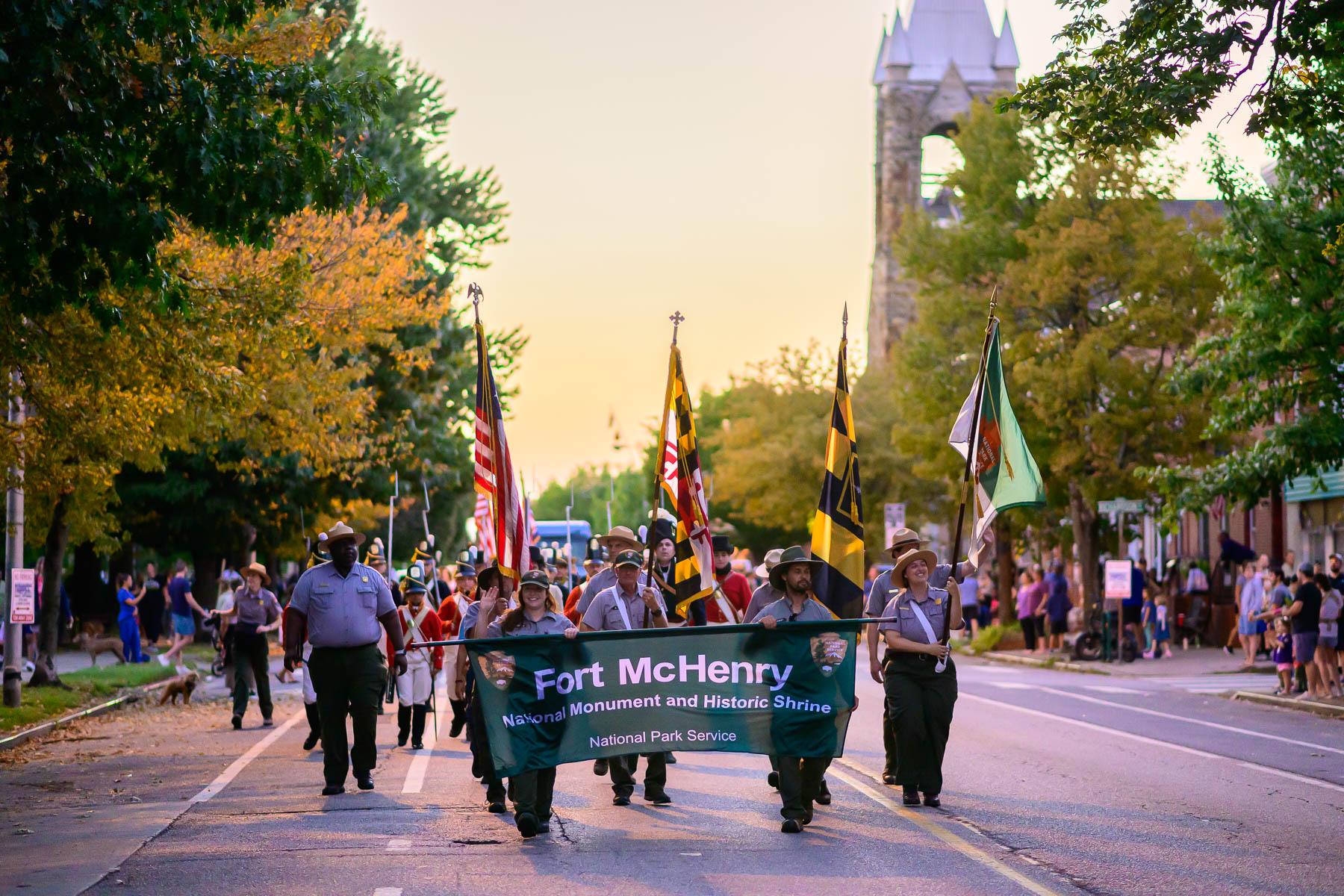 National Park Service rangers carrying flags and a Fort McHenry banner in front of a parade.