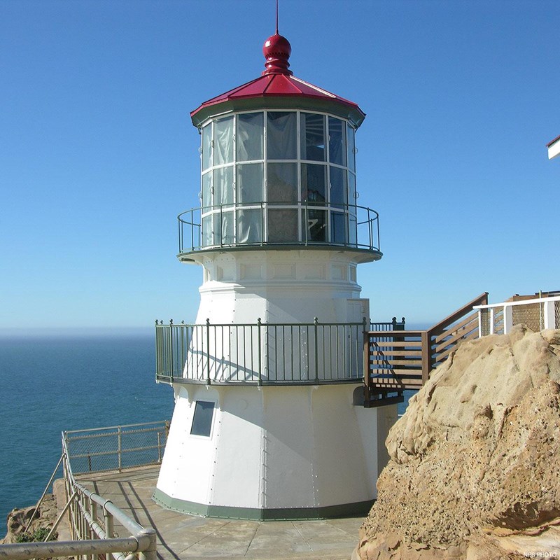 A three-story lighthouse with white sides and a red roof on the edge of an ocean-facing cliff.