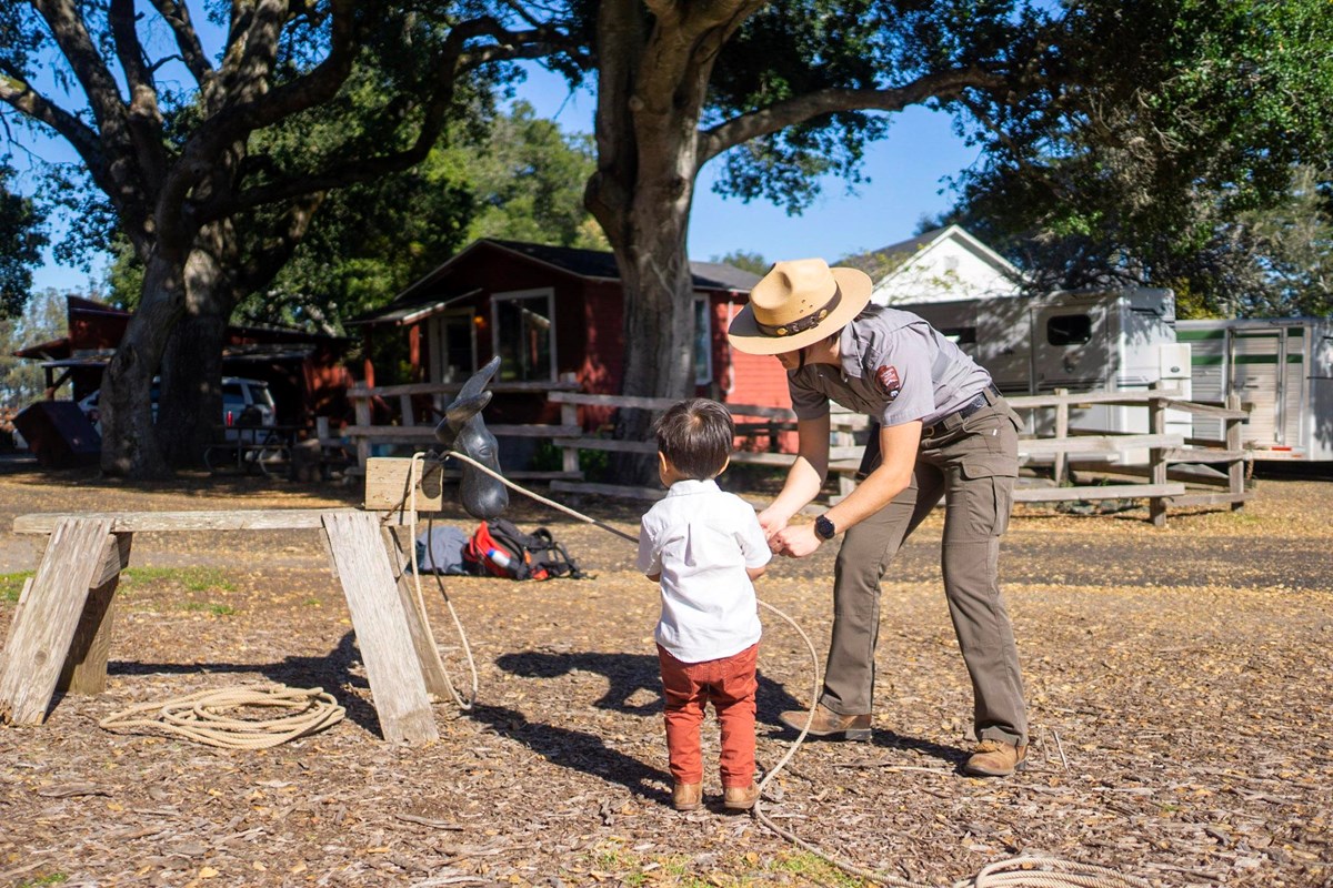 A park ranger helps a small boy handle a rope next to a small wooden calf dummy.