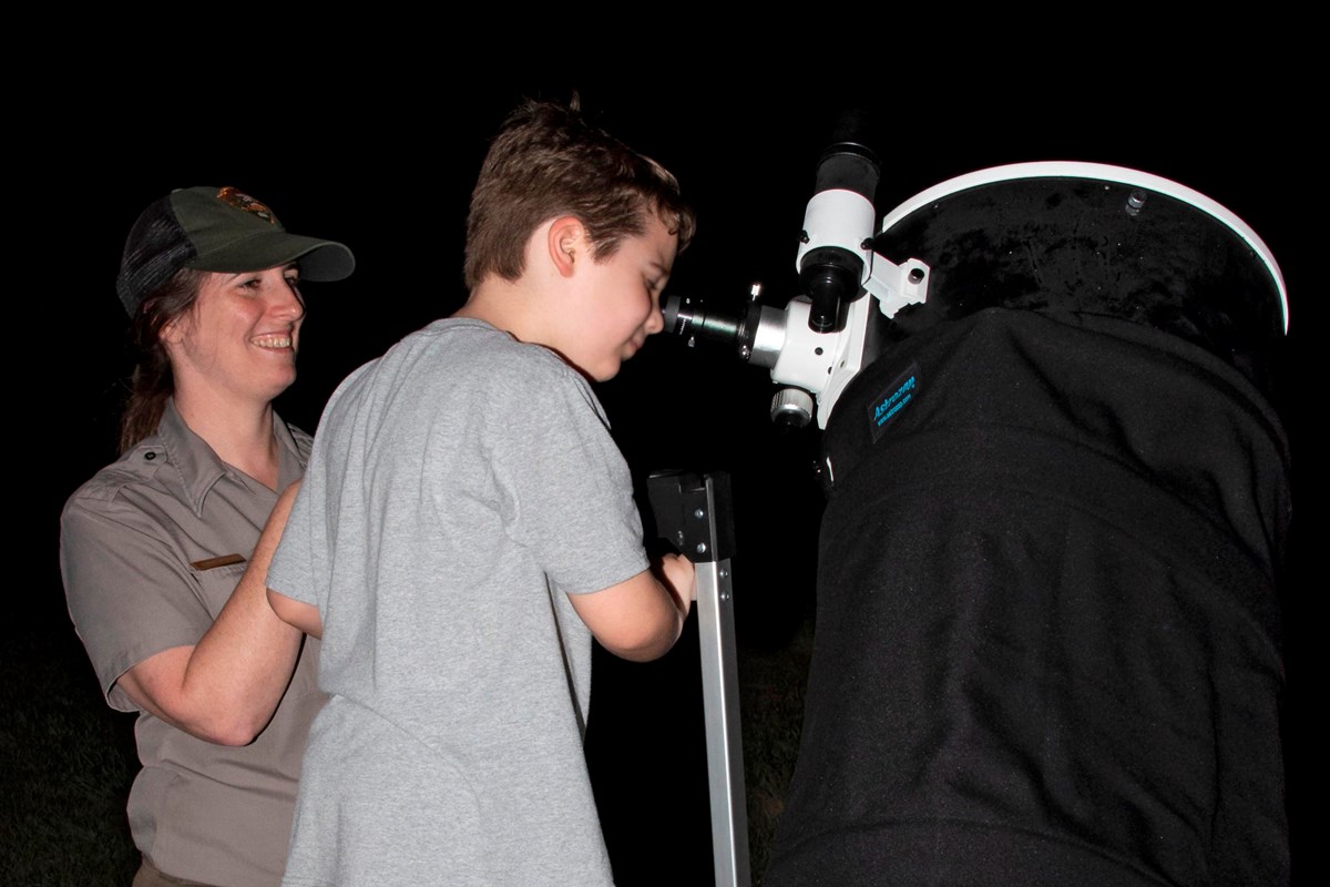 A young boy looks through a large telescope while a ranger looks on.