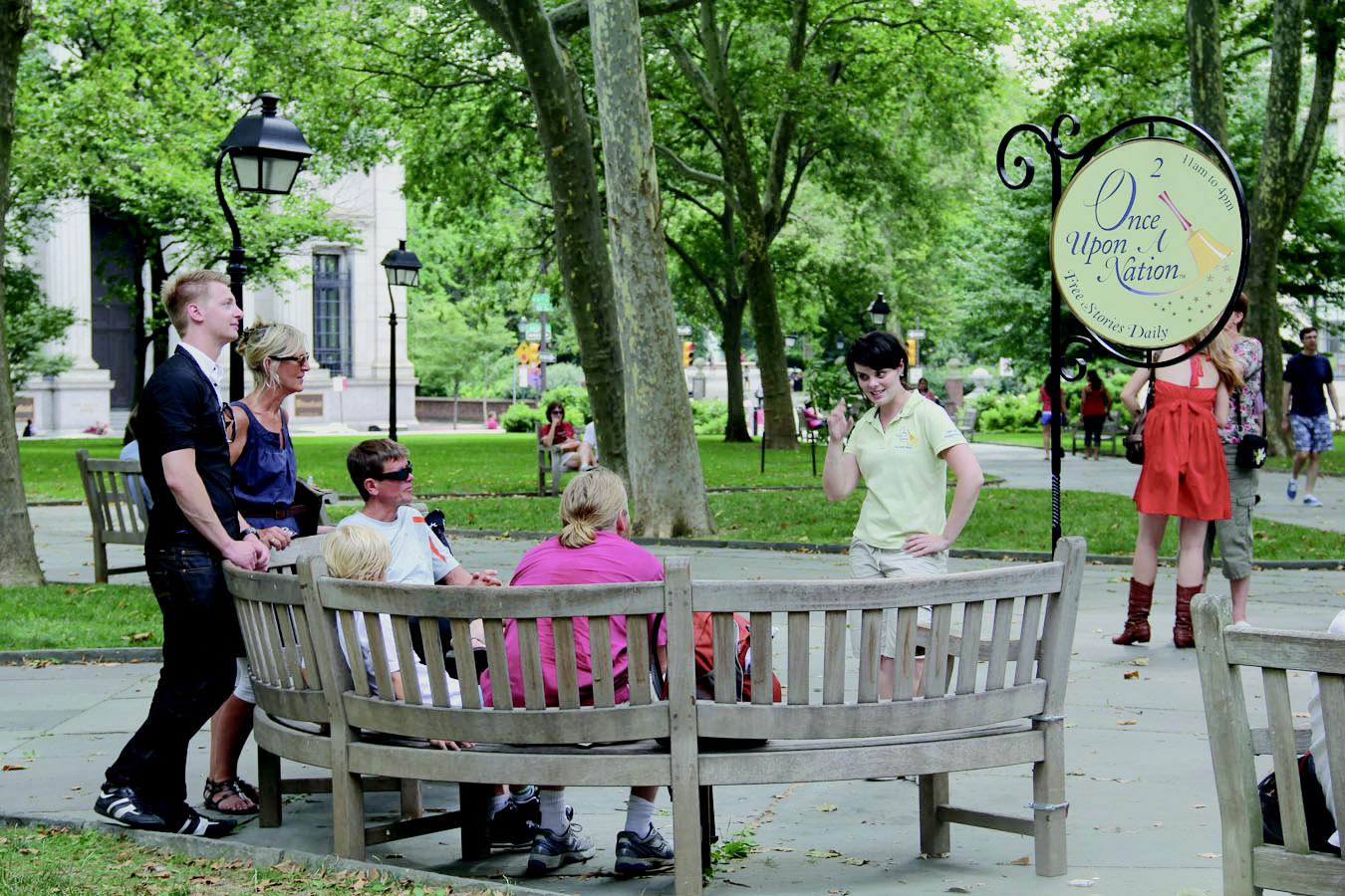 Visitors sit outdoors on a semi-circular bench, facing a person who is telling a story.