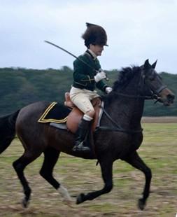 A white man wearing a British green coat and white trousers riding a horse.