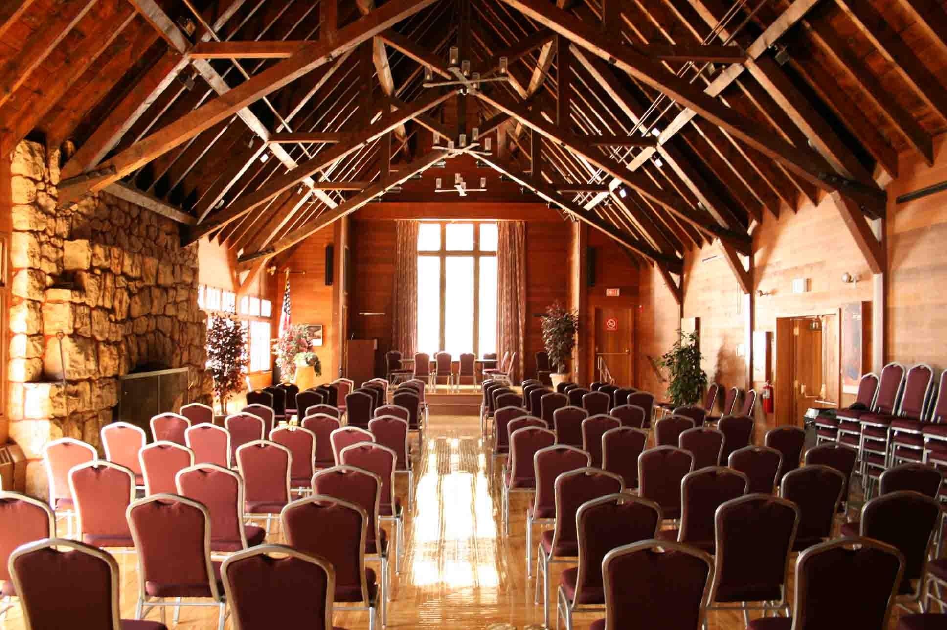 Large room with a lot of woodwork, a stage at one end, and multiple chairs facing the stage.