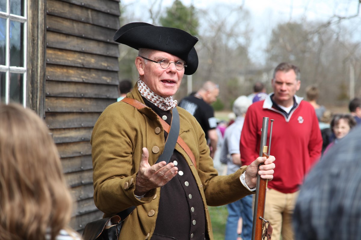 A man in a brown colonial coat and black colonial hat, carrying a musket, talks to modern visitors.