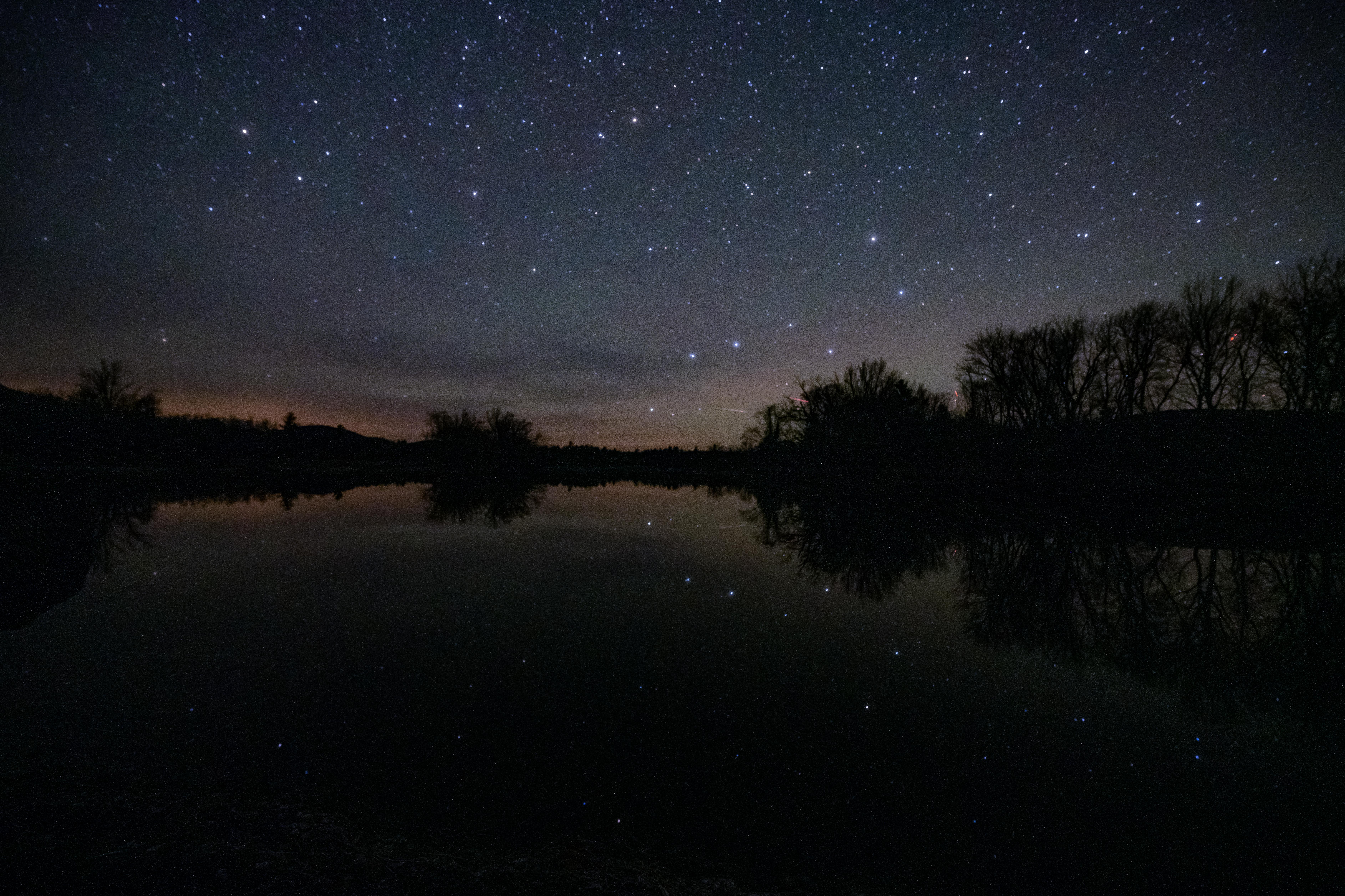A dark sky full of stars shows the big dipper, which is reflected on the tree-lined river below.
