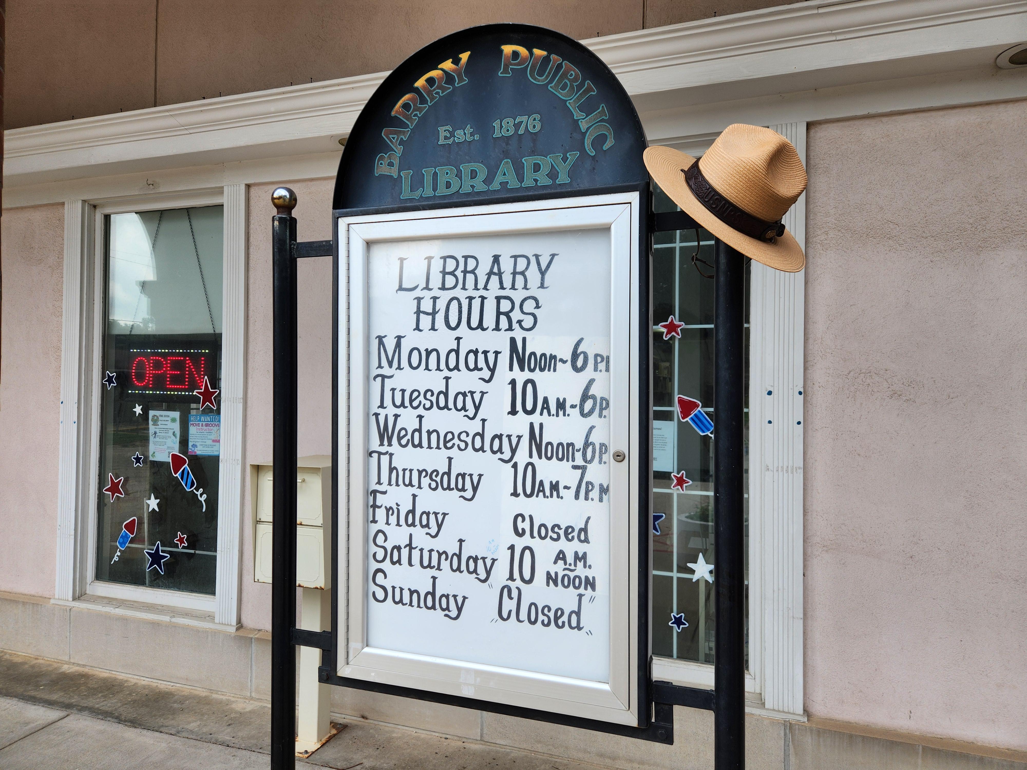 A ranger hat sits on top of a metal sign advertising the Barry Public Library's hours.