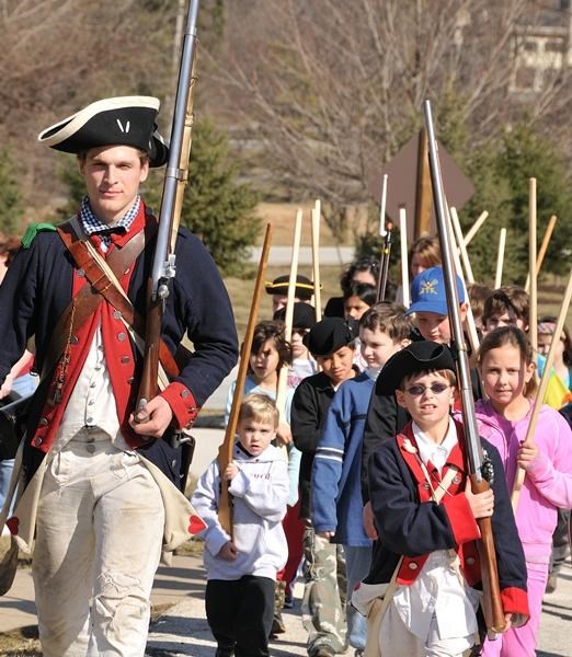 Park Ranger dressed as a Continental soldier marches children, who carry wooden training muskets