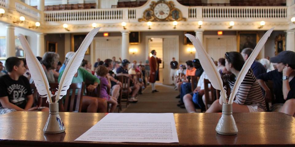 Quill pens on a desk flank a document on a table. In background an audience looks at a speaker.
