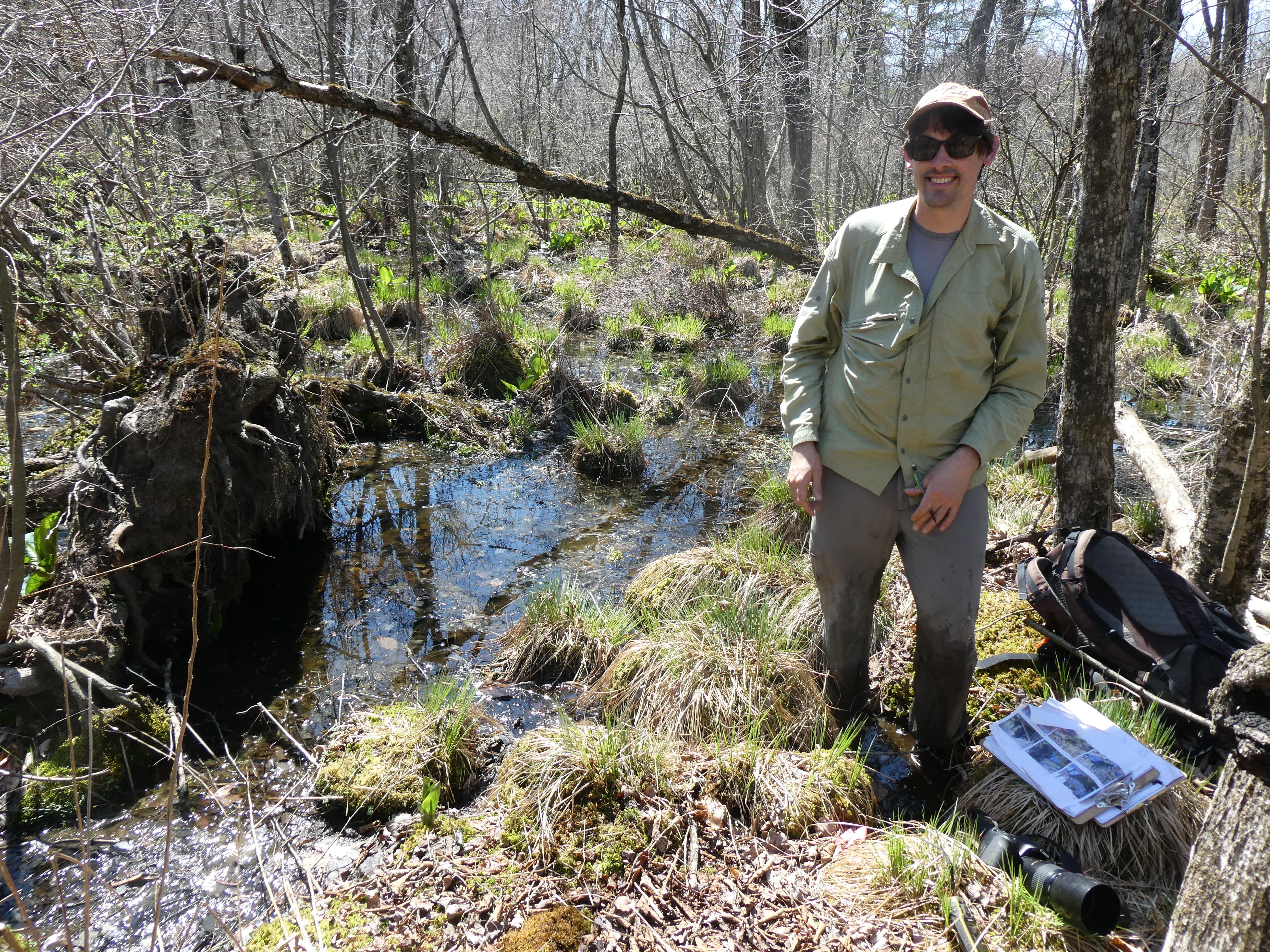 Person standing in a shallow wetland, surrounded by trees and grasses.