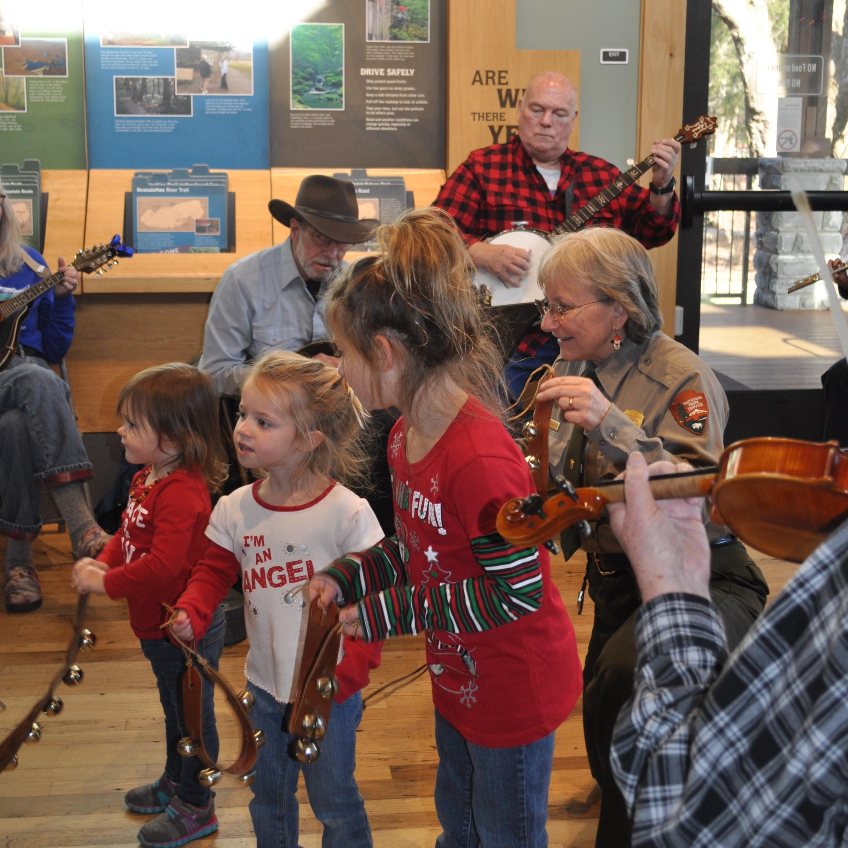 Three kids holding sleigh bells near a park ranger and people playing instruments.