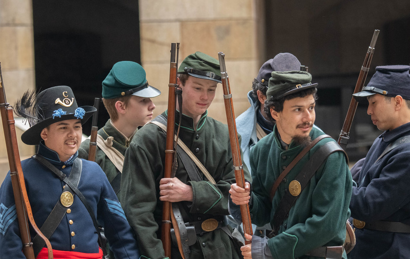 People dressed in soldier uniforms stand inside the fort holding rifles.