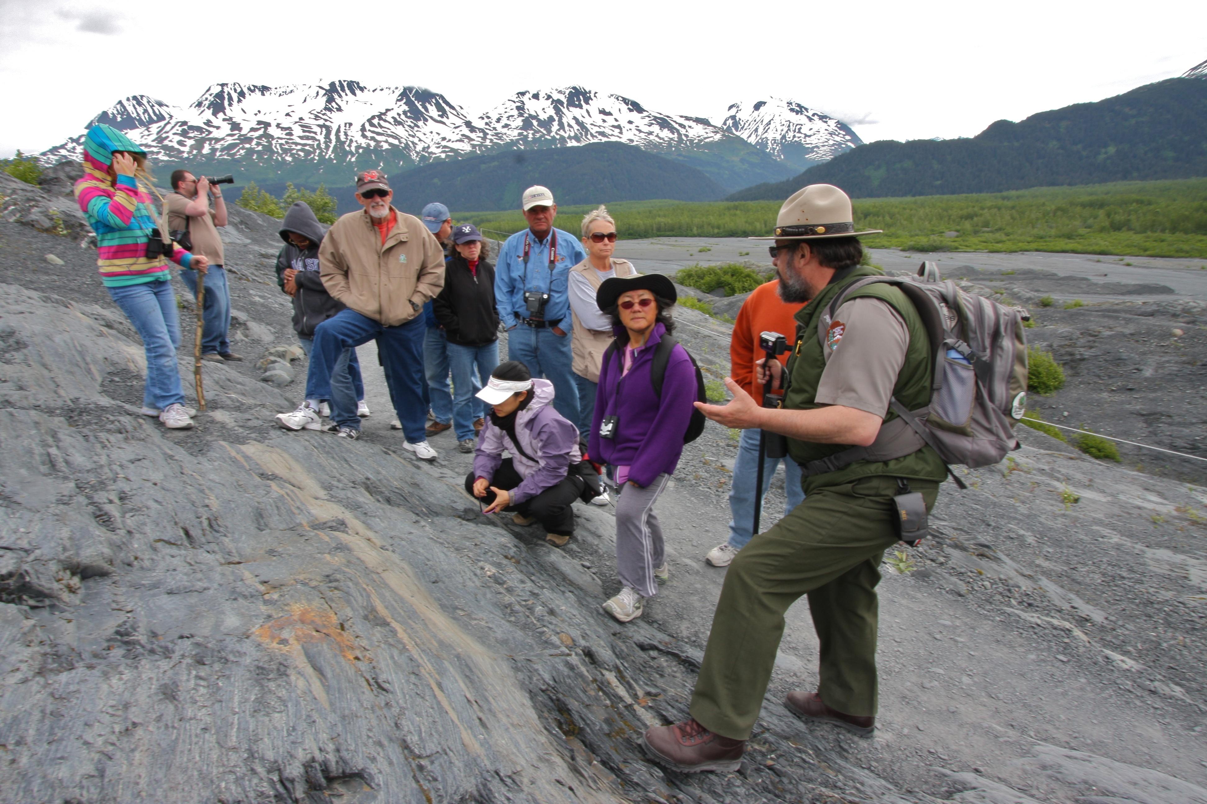 A ranger speaks to a crowd about geological features with snow-covered mountains in the distance.