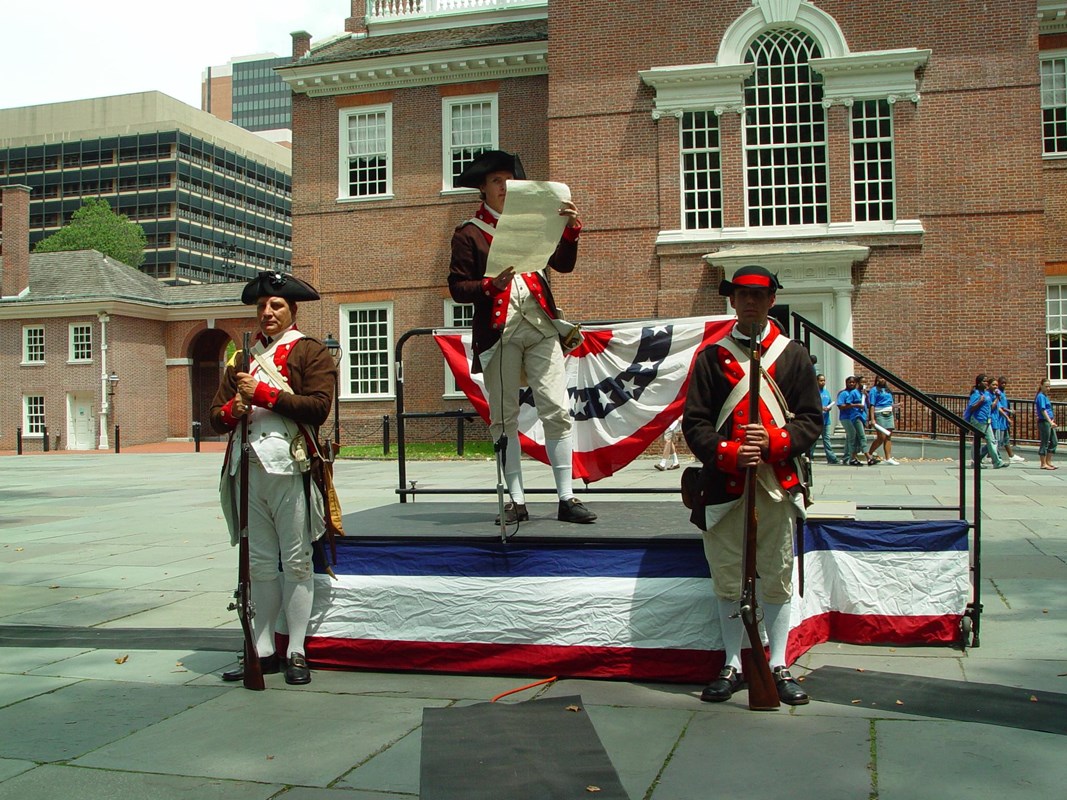 A man wearing a Revolutionary War uniform reads a scrolled document from a raised platform.