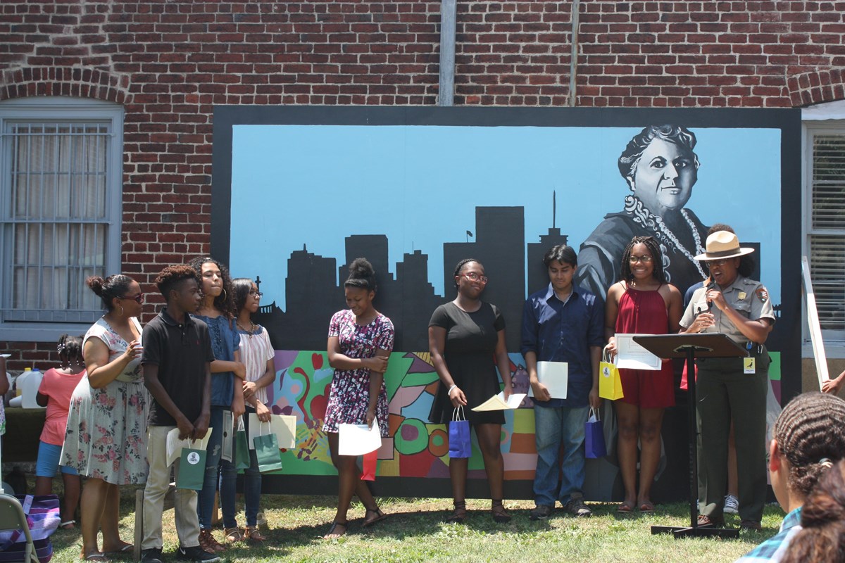 Ranger with students in front of a mural of Maggie L. Walker