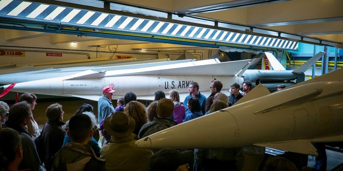 A crowd of about 20 visitors huddles around a Nike volunteer standing in front of a large missile