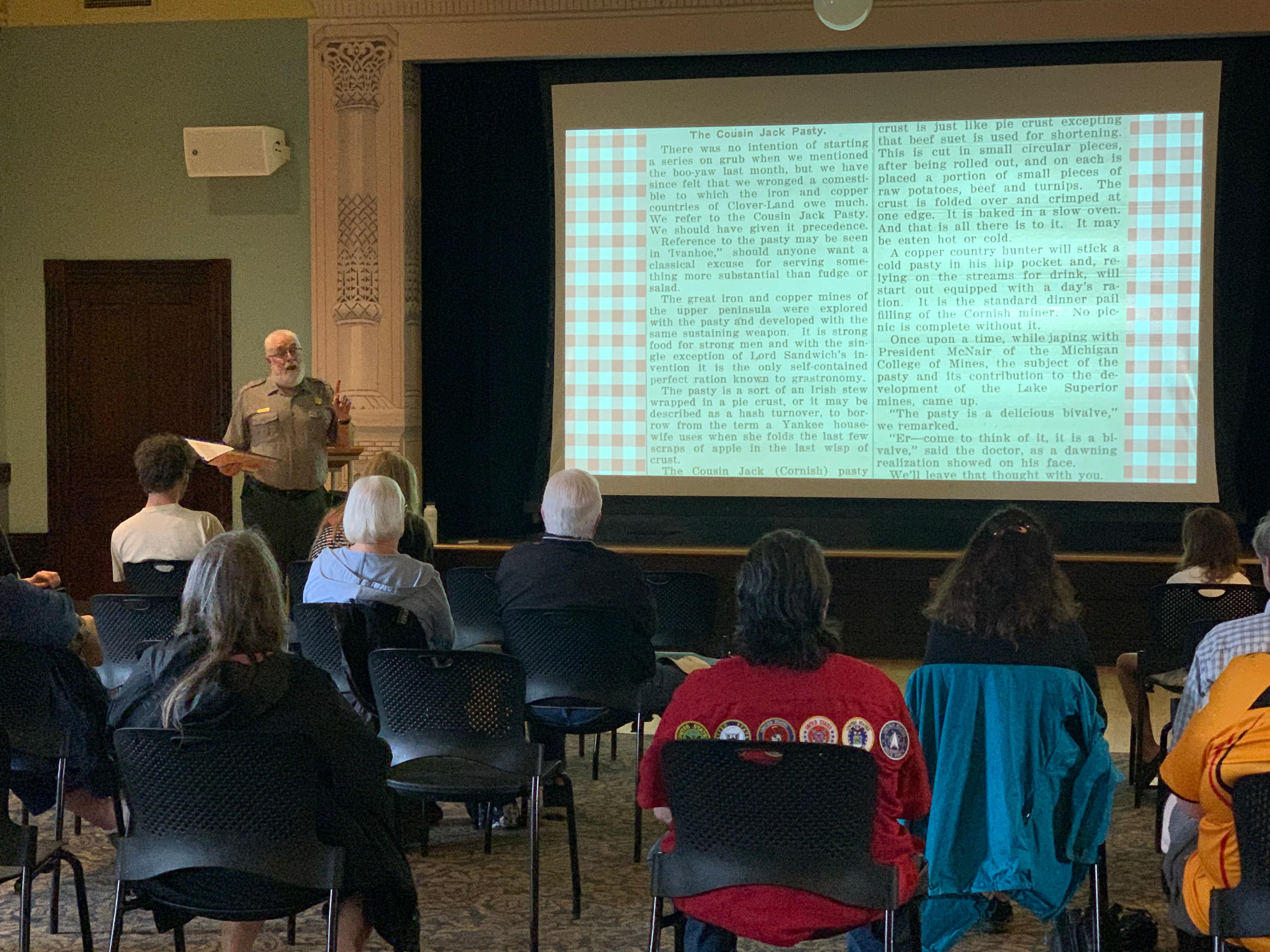 Visitors sit listening to a ranger give a presentation with a large image projected on a screen.