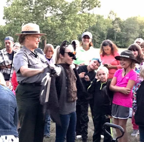 Park ranger engaging a group of children and adults during an outdoor educational event in a wooded
