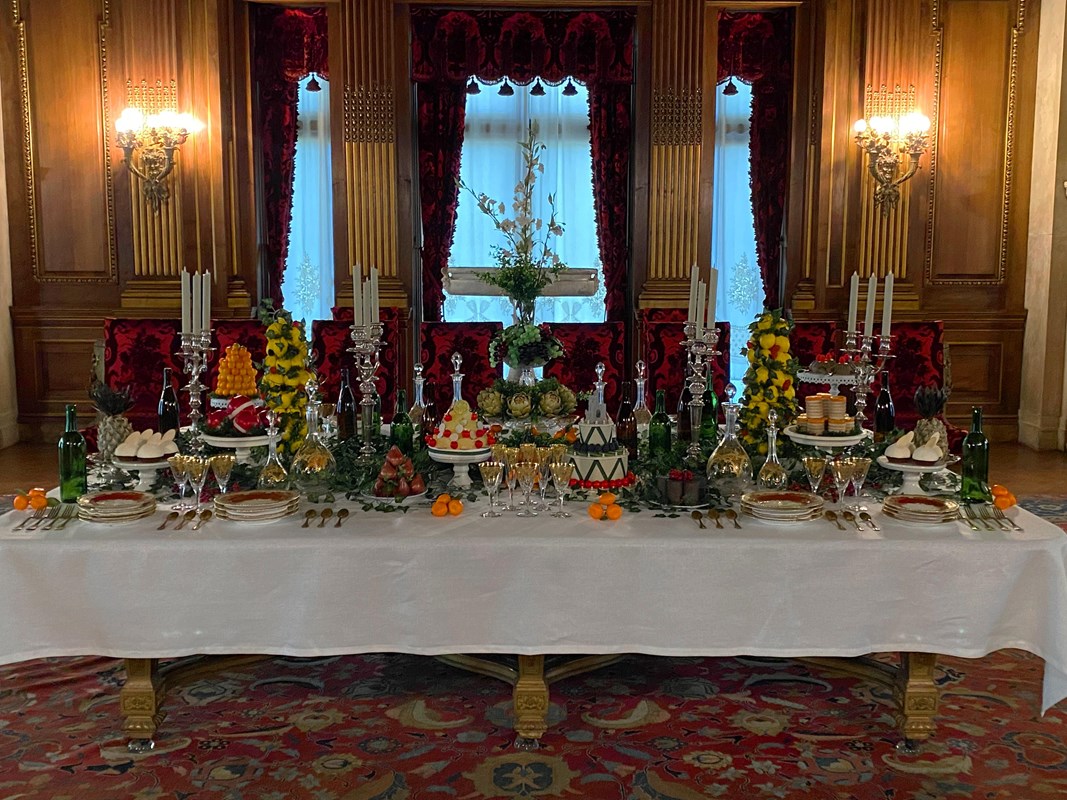 A long table in a large opulent dining room set with china and extravagant desserts.