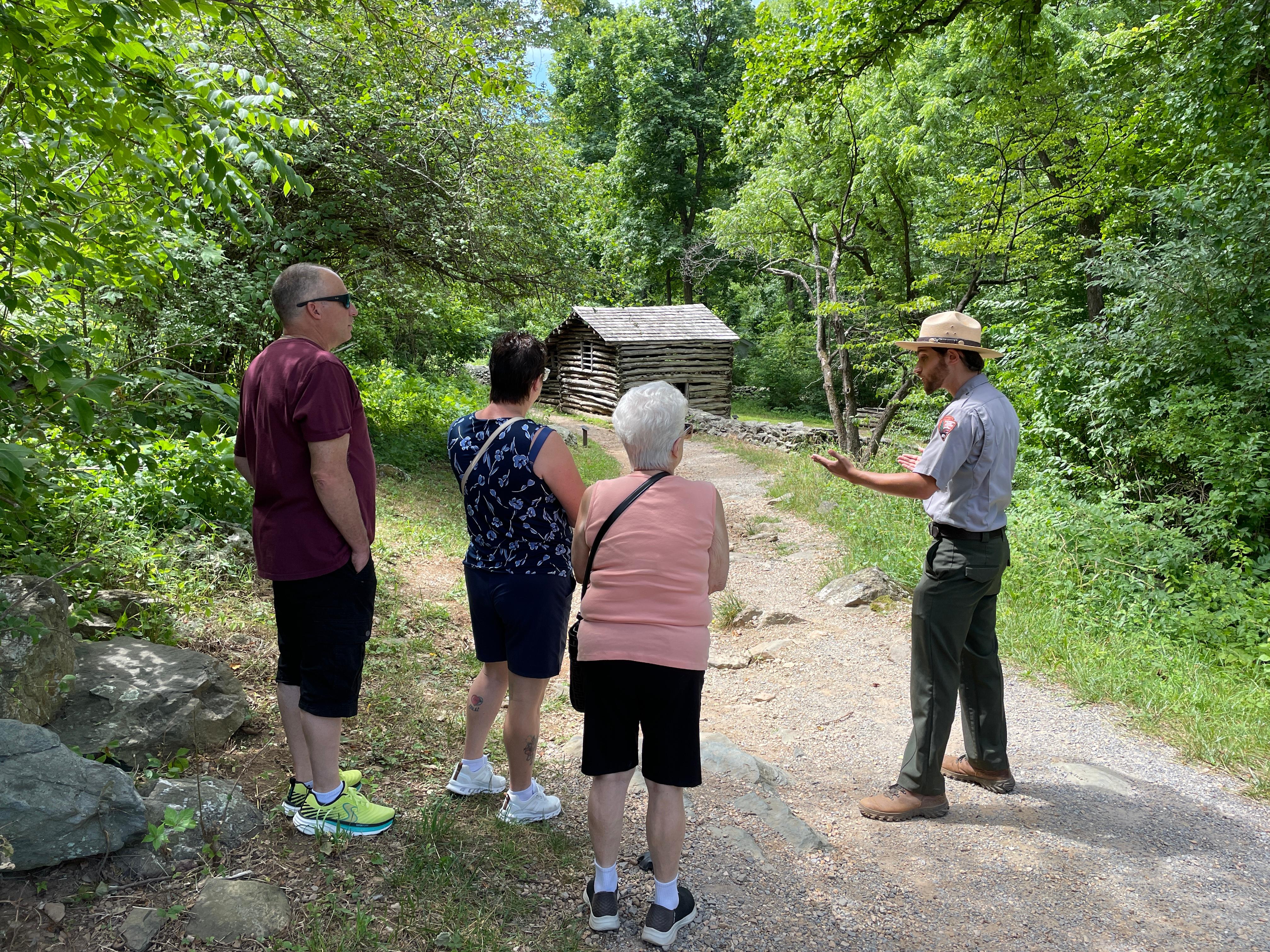 3 adults stand outside with a uniformed ranger on a dirt path with a log cabin in background