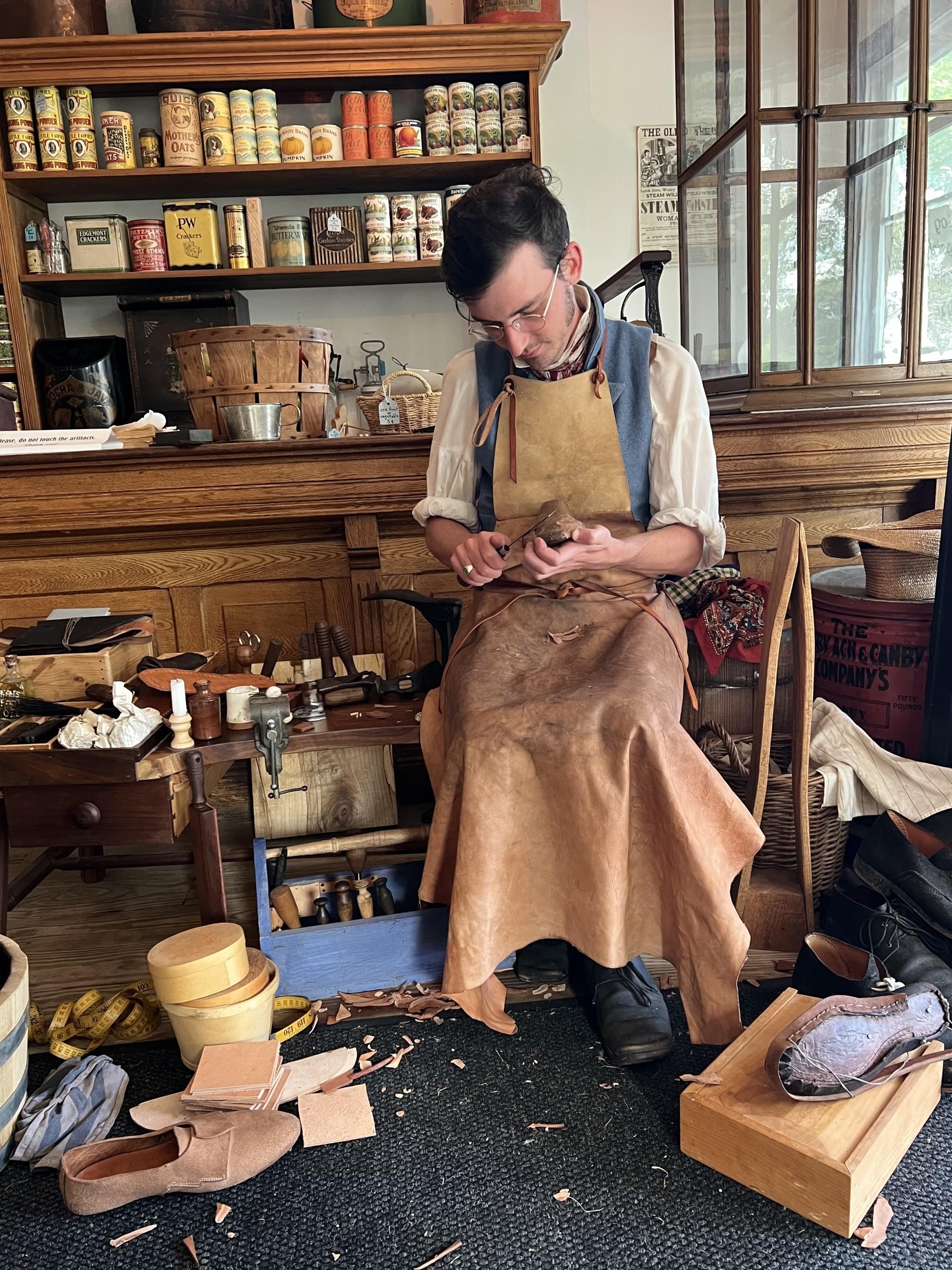 A living history interpreter in period-accurate 19th-century attire sits in a wooden workshop while