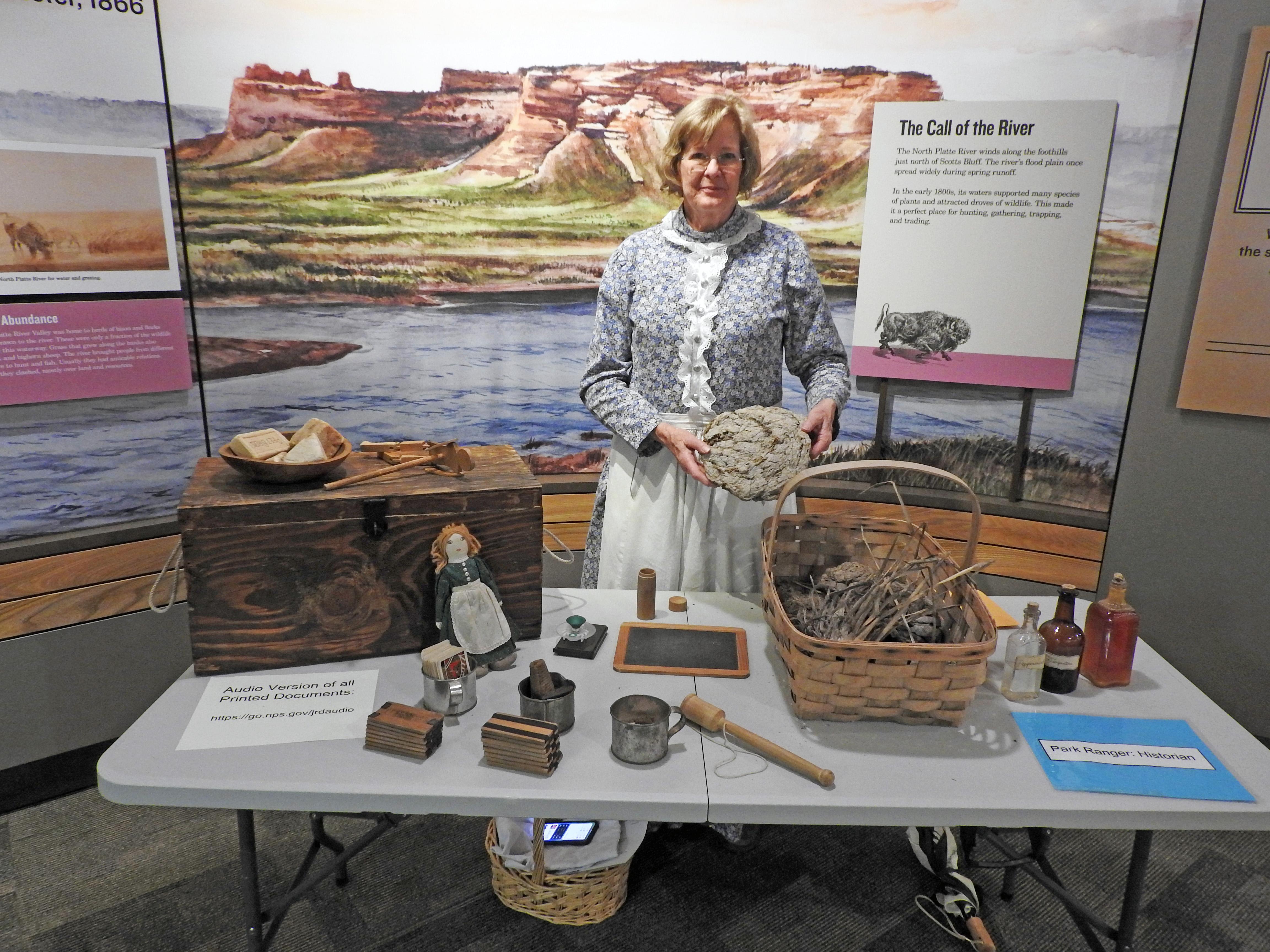 A woman is dressed in a calico dress and standing behind a table that is covered with old time toys.