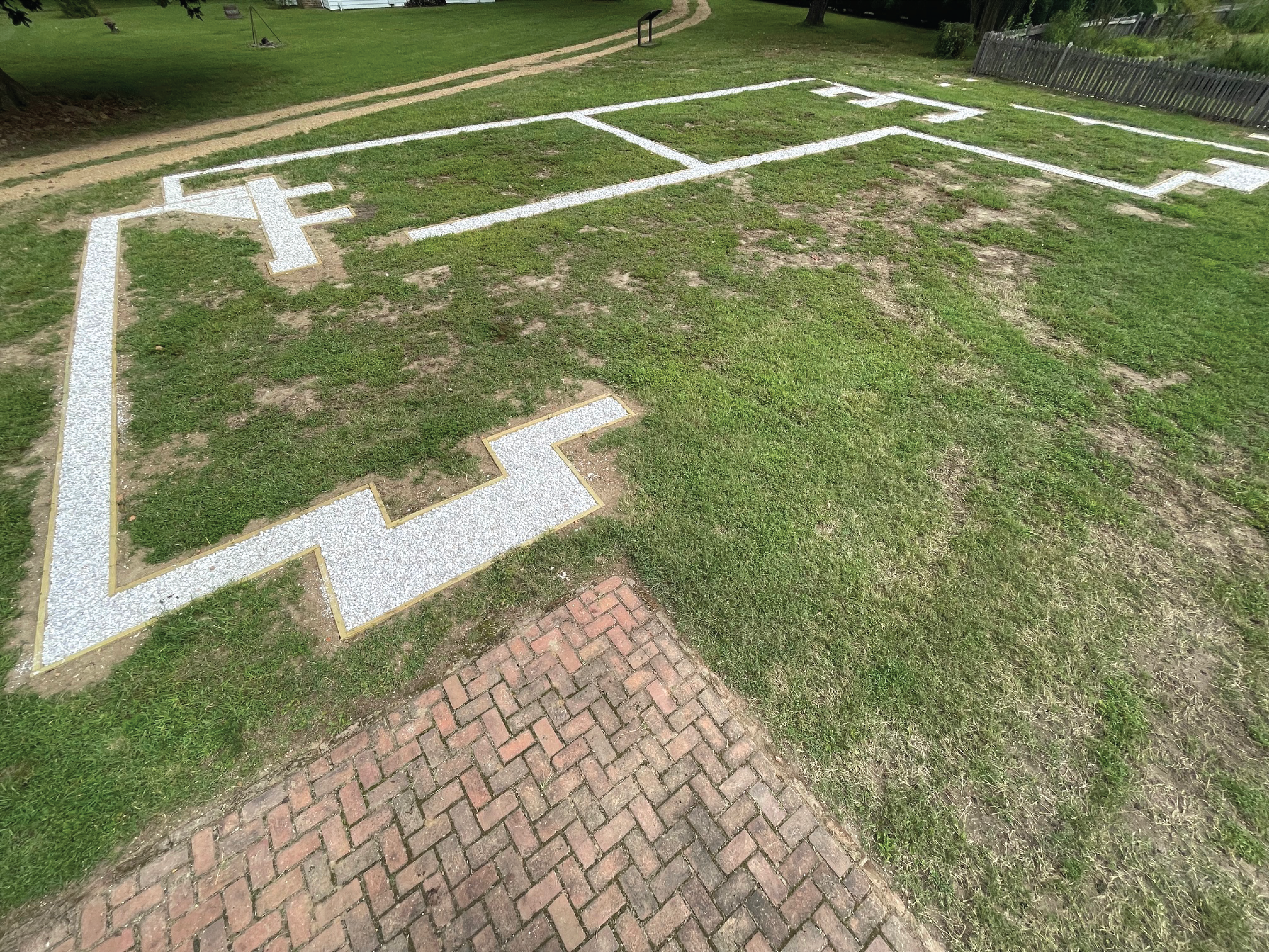 A grassy field with a white stone outline of a building, with a brick patio in the foreground.