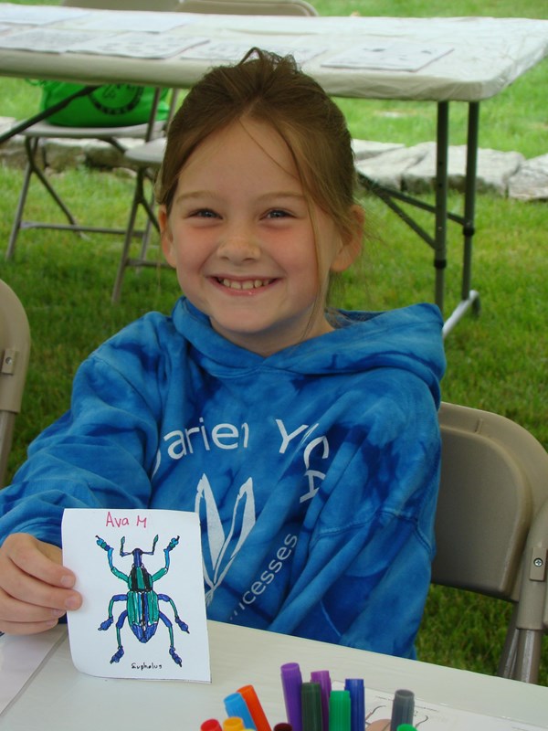 A young girl with a blue sweatshirt on sits at table and holds her artwork.