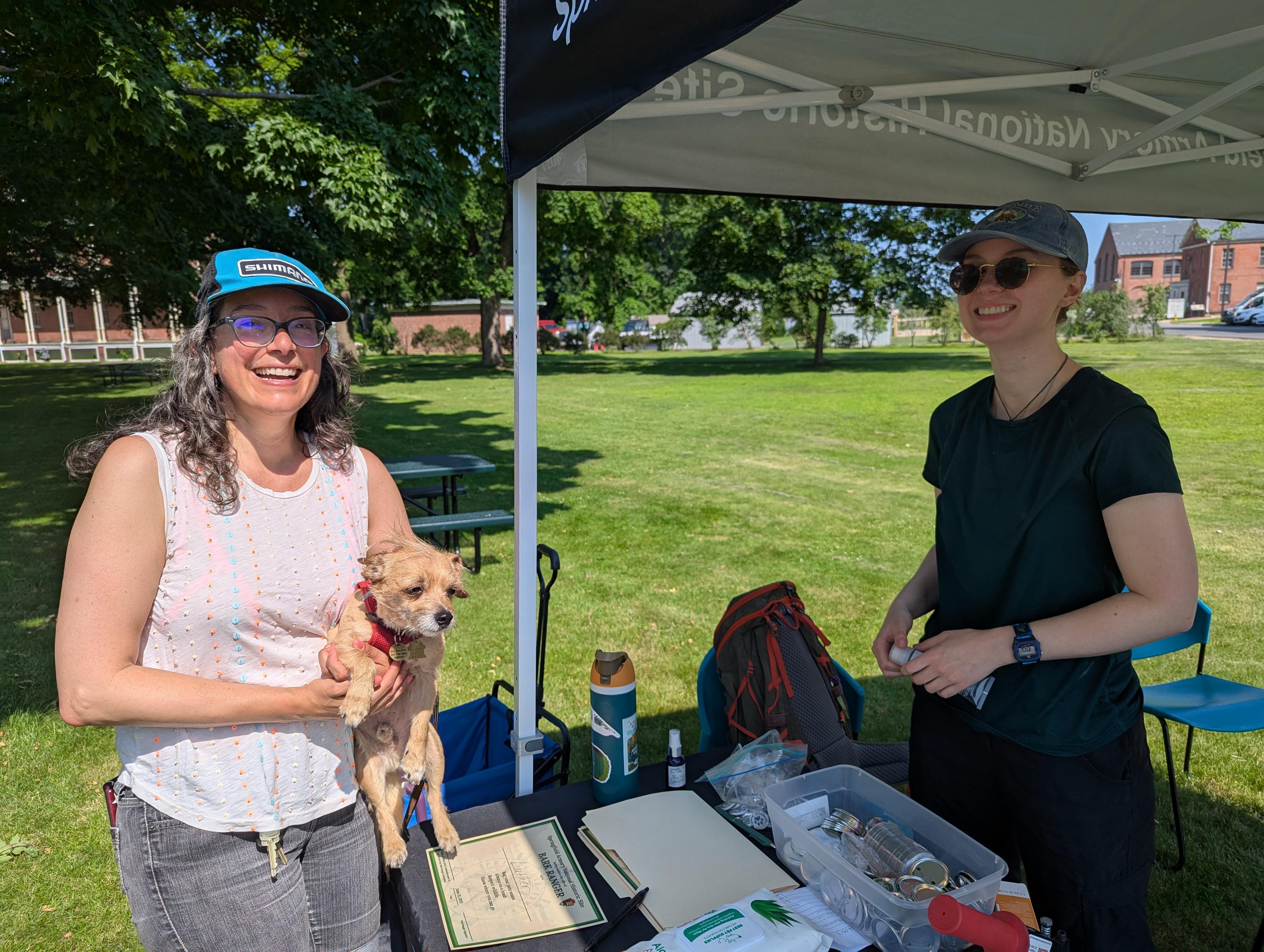 Two women face the camera.  One holds a small dog.  The other is under a tent awning.