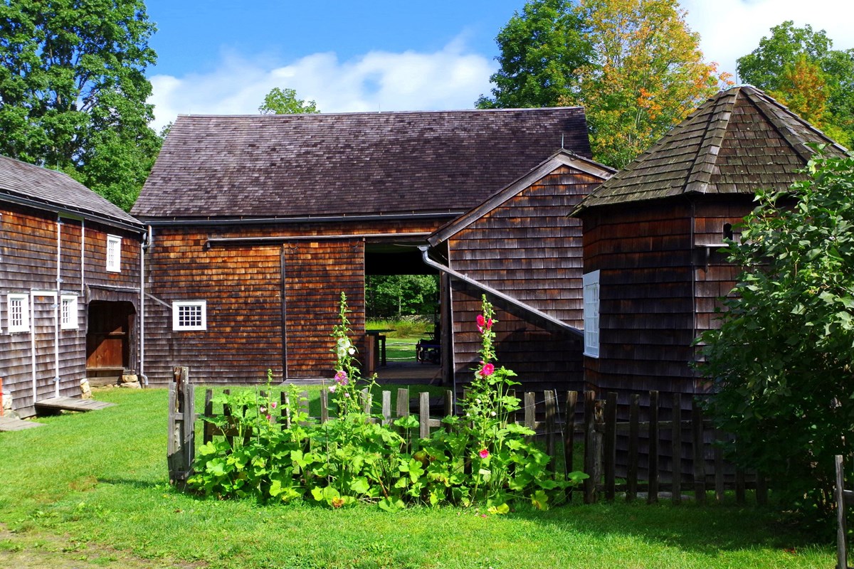 A brown wooden barn with a wooden fence and flowers in front of it.