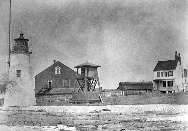 A light house and guard tower stand before two historic buildings set in black and white.