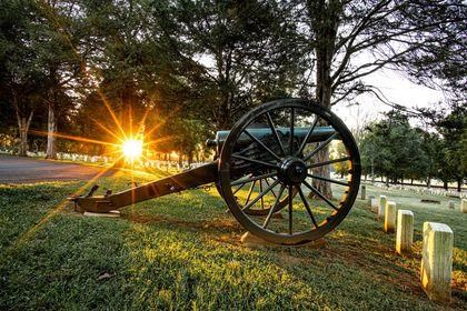 The sun rise is visible through trees shining on rows of white headstones and a Civil War era cannon