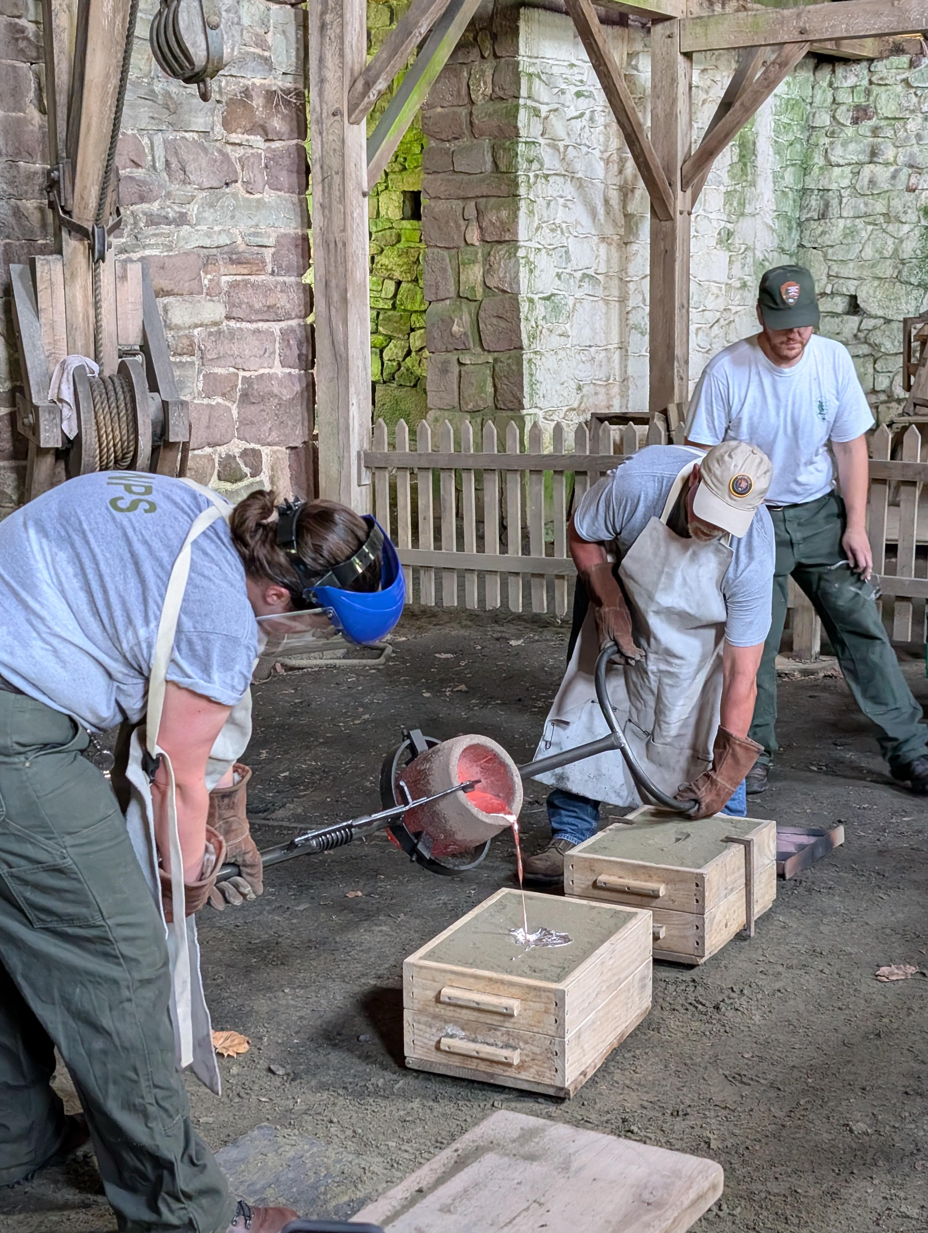 Two people pour molten aluminum from a crucible into sand molds on the ground.