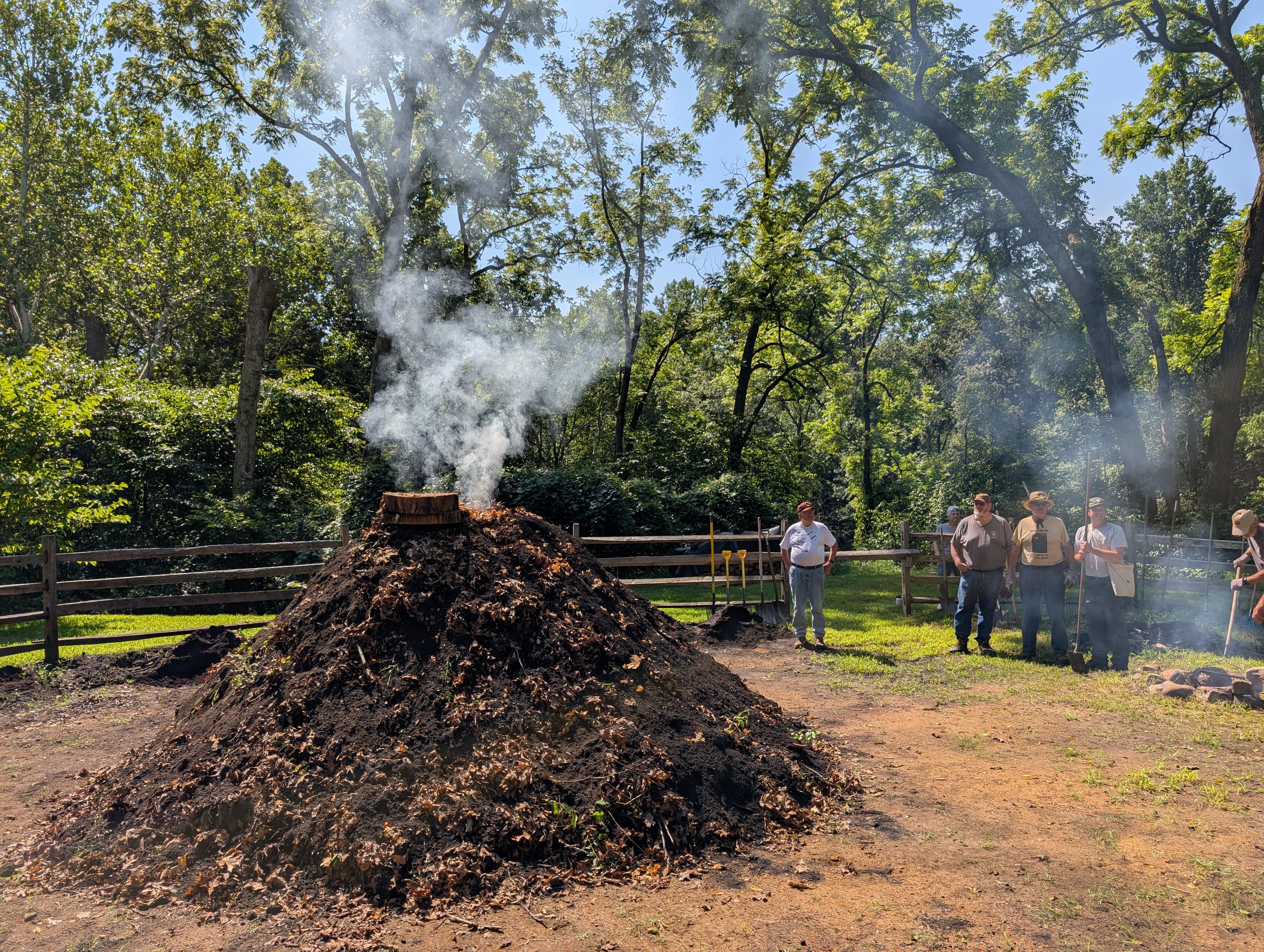 A dirt covered stack of wood emits smoke from the top while a group of people watch.