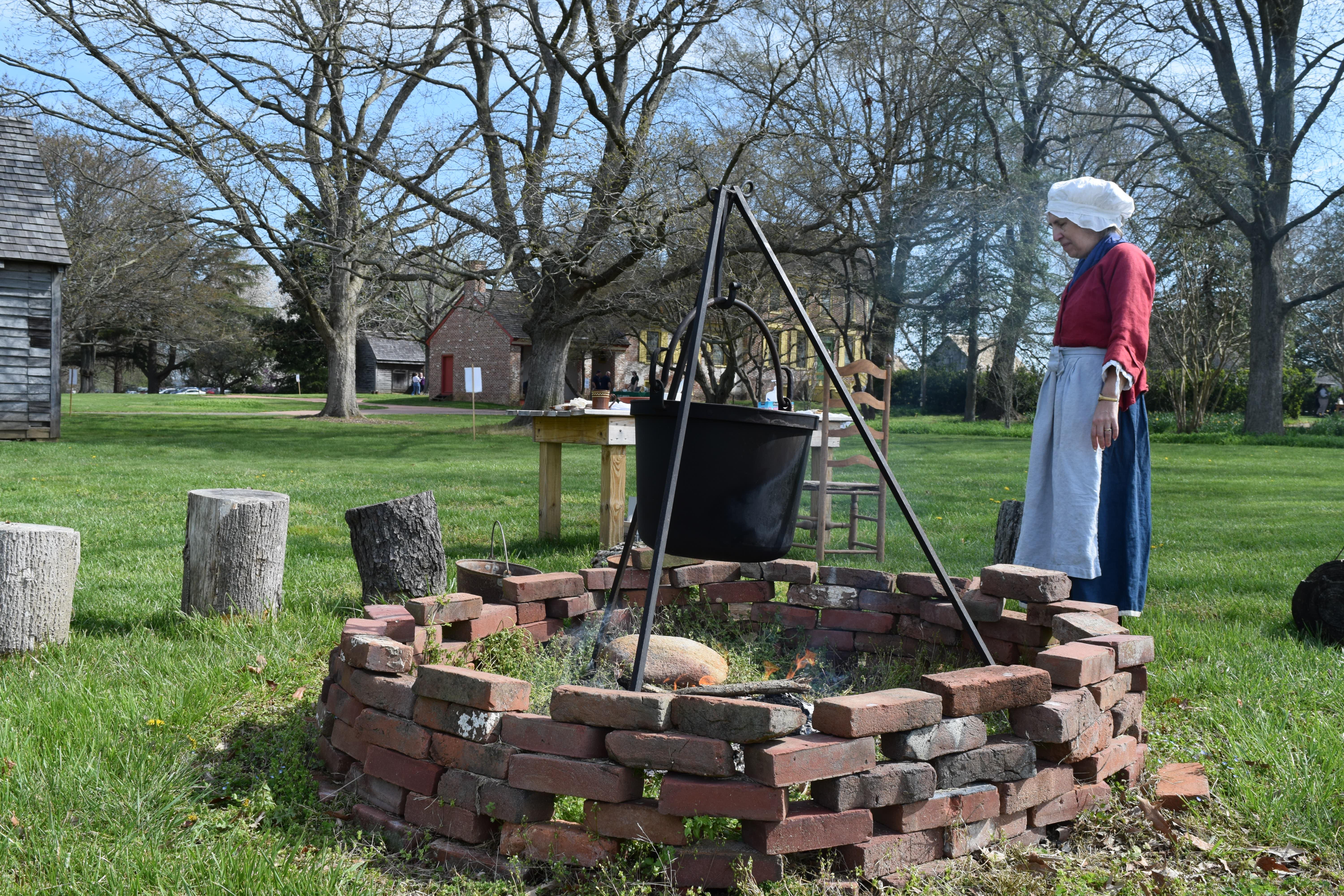 A women dressed in historical clothing cooks over a fire.