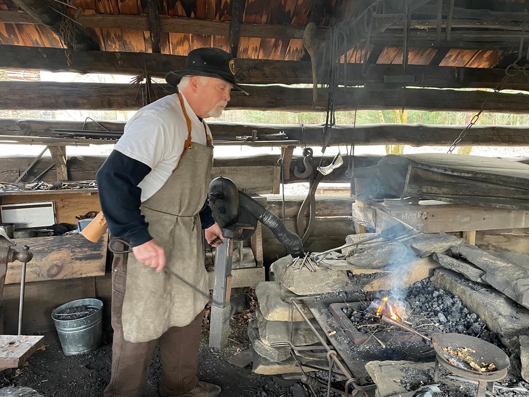 A standing in a cabin-like structure wearing a work apron while tending to a fire to mold metal.