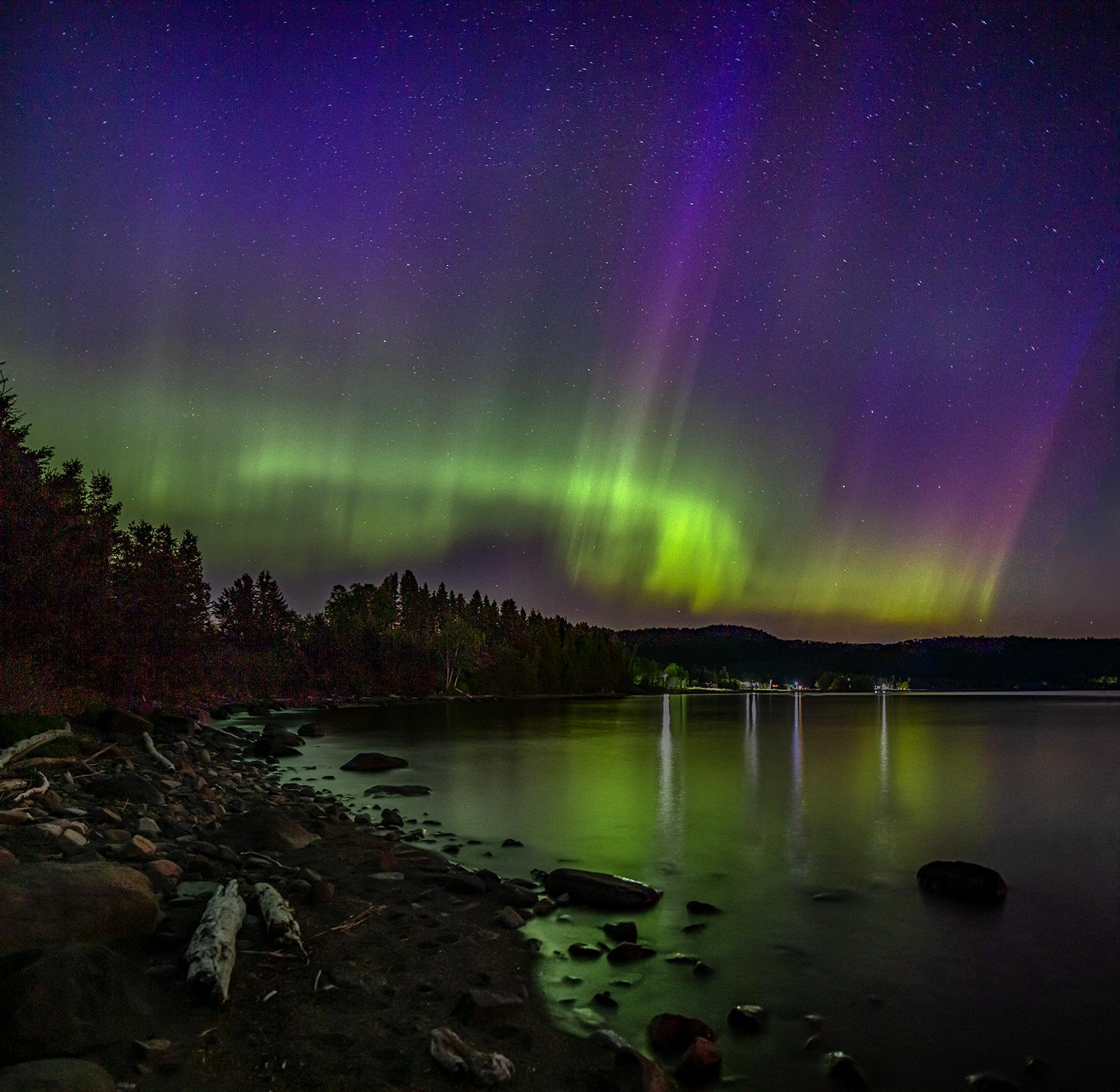 Green-streaked night sky over a bay with reflections of lights along the shore.