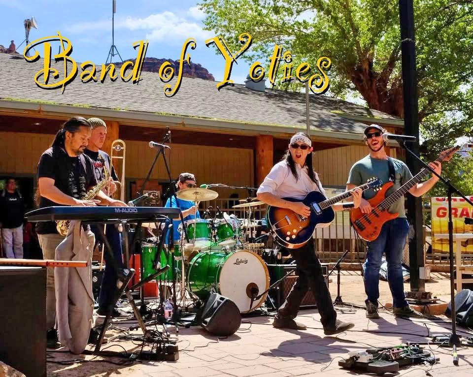 Five musicians in a rock band with guitars, a keyboard and drum set are performing outdoors.