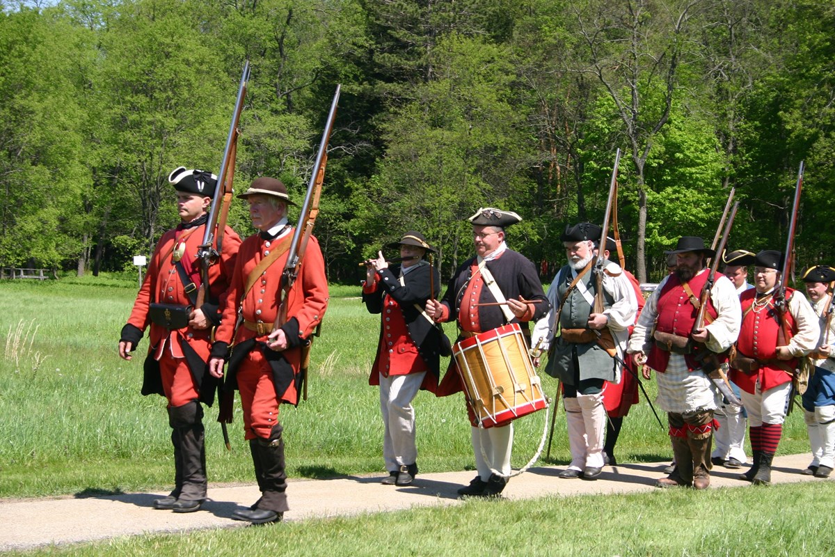 Re-enactors in red military uniforms and carrying flintlock muskets march along a path in a meadow.