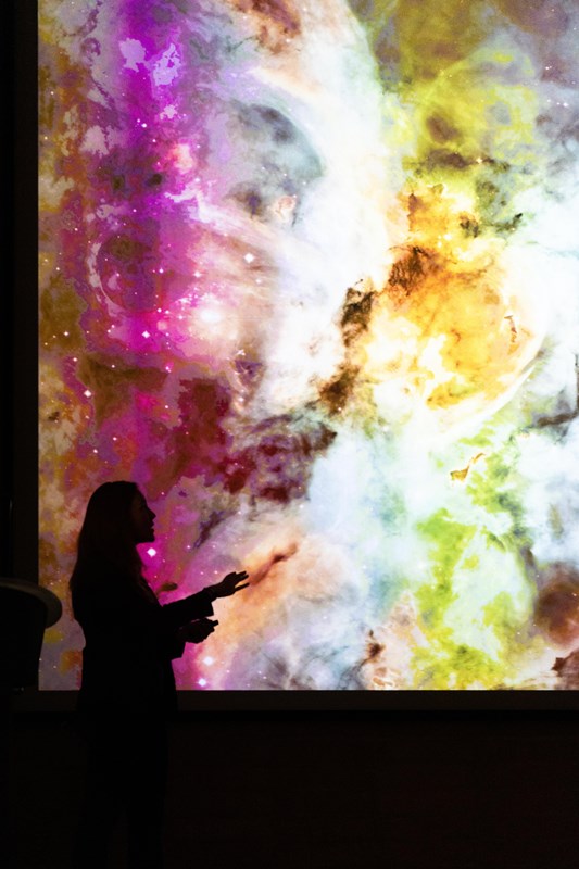 Silhouette of a person on stage in front of a colorful photo of a nebula on a PowerPoint slide.