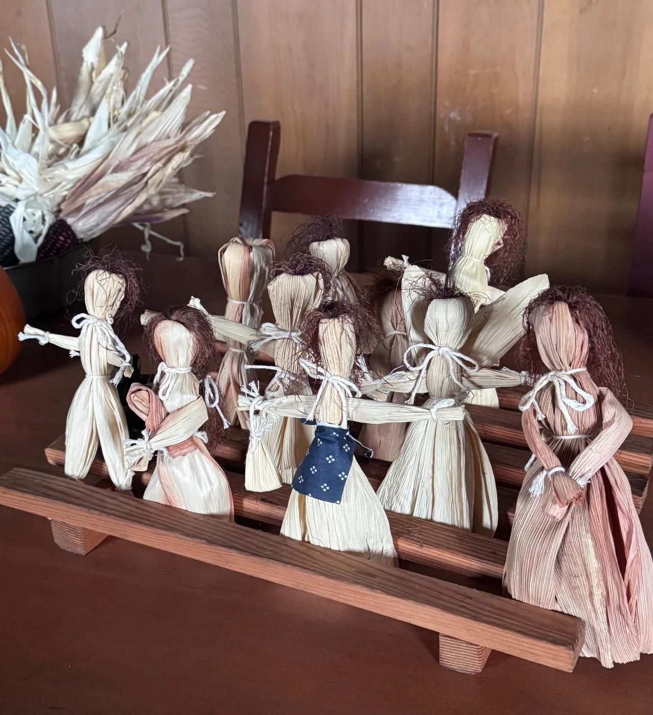A group of handmade corn husk dolls displayed on a wooden rack, each tied with string and some decor