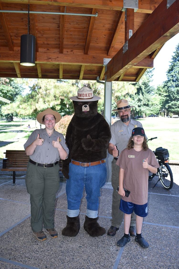 Smokey Bear stands between two National Park Service rangers and a child under a pavilion, all smili