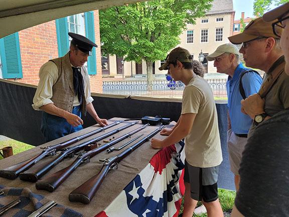 A man wearing clothing from the early 1800s displays a table of weapons for a group of visitors.